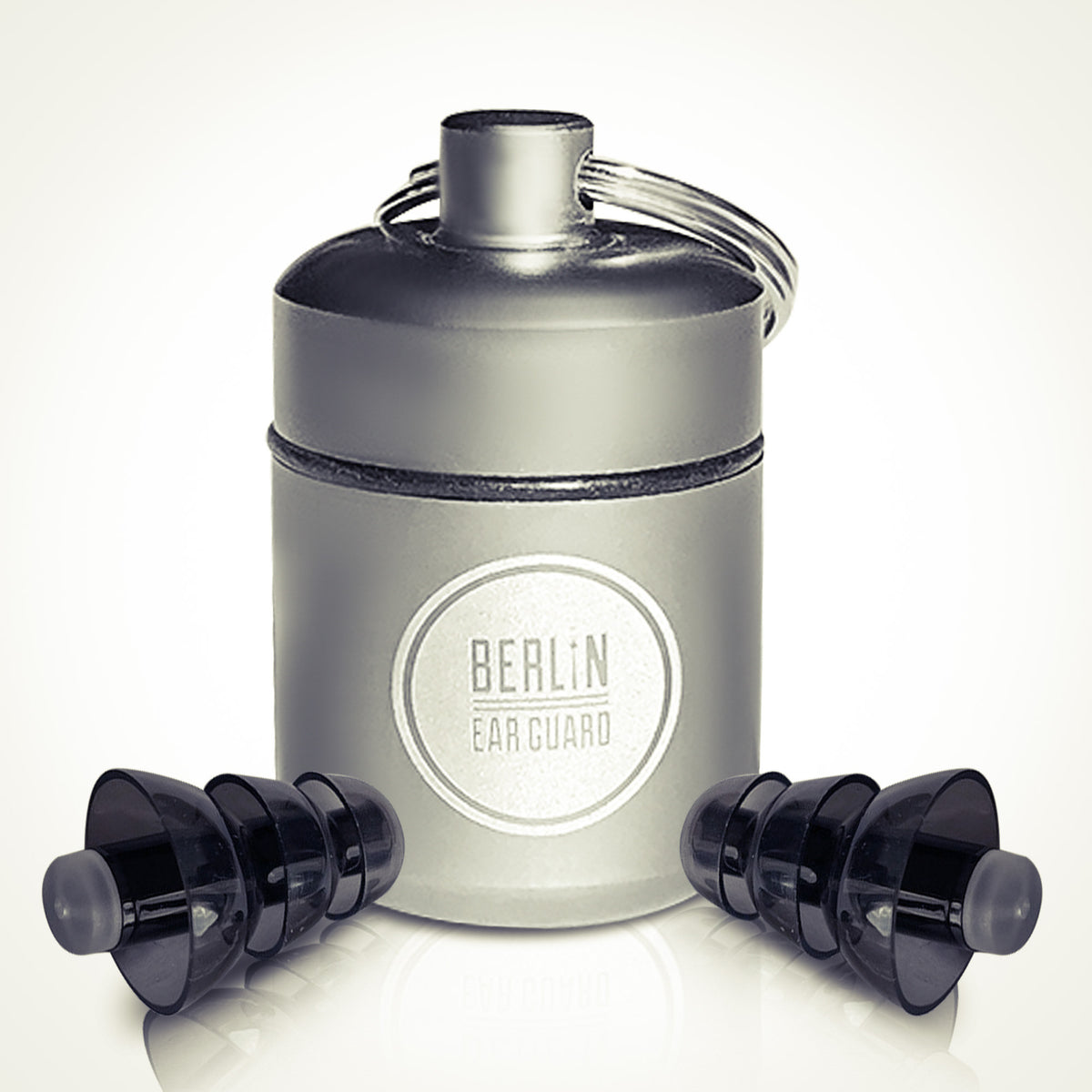 Ein silberner, zylindrischer Berlin Ear Guard-Behälter mit einem Schlüsselring, flankiert von zwei schwarzen Silikon-Ohrstöpseln, einer auf jeder Seite, vor einem hellen Hintergrund.